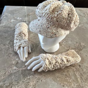 May include: A cream-colored crochet hat and matching fingerless gloves set. The hat has a rounded crown and a small brim. The gloves are designed to cover the hand up to the knuckles. The set is displayed on white mannequin hands.