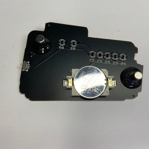 May include: A black circuit board with a silver battery, two black knobs, and a small white connector. The battery is labeled "Sunbeam 3V CR2032".