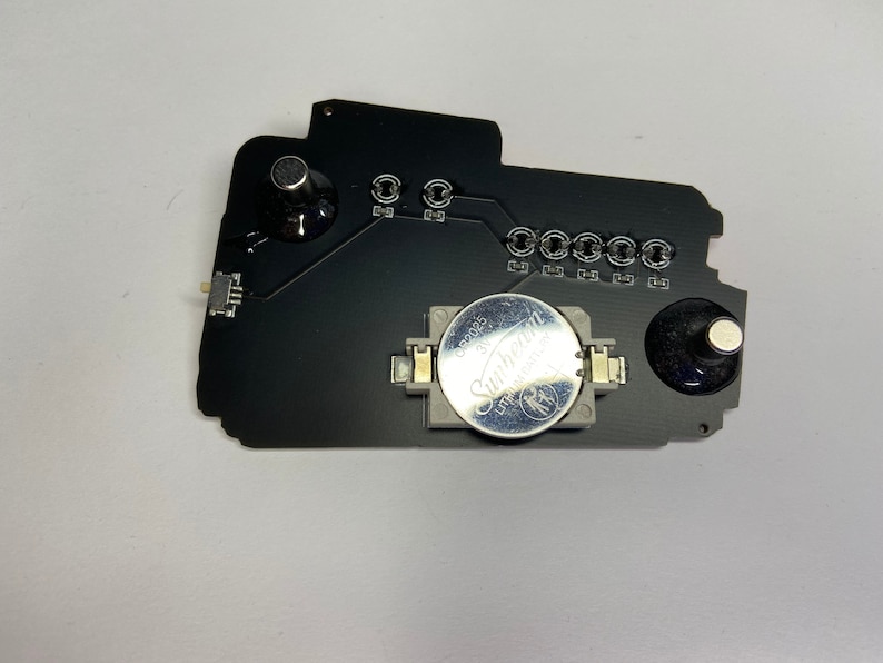May include: A black circuit board with a silver battery, two black knobs, and a small white connector. The battery is labeled "Sunbeam 3V CR2032".