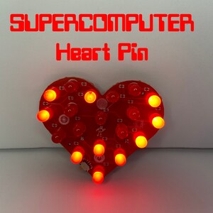 Puede incluir: Pin electrónico rojo en forma de corazón con LEDs iluminados. El texto "SUPERCOMPUTER Heart Pin" se muestra en rojo. Los LEDs del pin emiten un brillo cálido. Es probable que sea un accesorio portátil o un elemento decorativo.