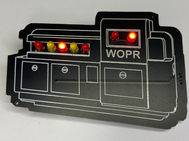 May include: A black WOPR computer with a white outline and red and yellow lights. The computer has a screen with two red lights on top and the word "WOPR" printed below.