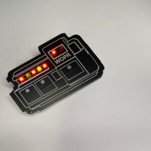 May include: Black WOPR computer system pin with a row of five red and yellow lights.