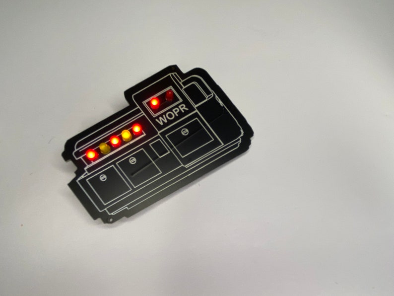 May include: Black WOPR computer system pin with a row of five red and yellow lights.