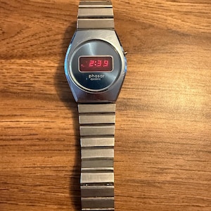 May include: A silver-tone digital wristwatch with a metallic link band. The watch face features a circular blue border surrounding a rectangular digital display showing the red LED time 2:39. The text "phasar" and "quartz" is printed below the display.
