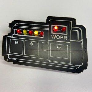 May include: A black WOPR computer circuit board with five red, yellow, and orange LED lights. The lights are lit up. The board has a white outline of a computer with the text "WOPR" printed on it.