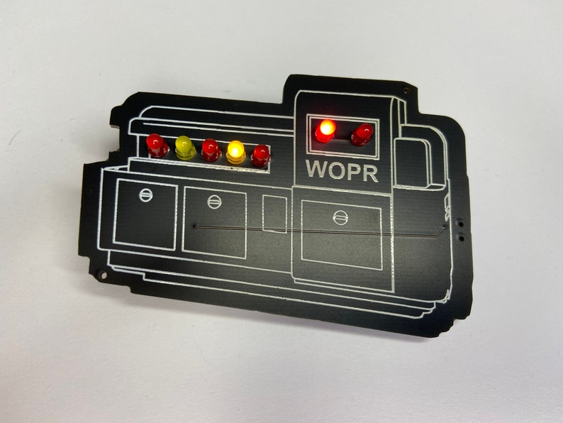 May include: A black WOPR computer circuit board with five red, yellow, and orange LED lights. The lights are lit up. The board has a white outline of a computer with the text "WOPR" printed on it.