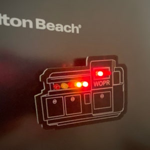 May include: A black and white illustration of a machine with red and yellow lights. The machine has a label that says "WOPR".