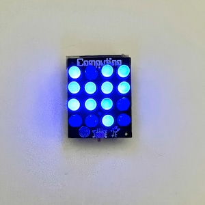 Può includere: Un pannello rettangolare nero con la parola "Computing" stampata su di esso. Il pannello ha 16 luci LED blu disposte in una griglia 4x4.