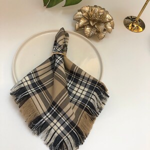 May include: A brown and black plaid napkin with fringed edges, folded and secured with a gold ring. The napkin is placed on a white plate.
