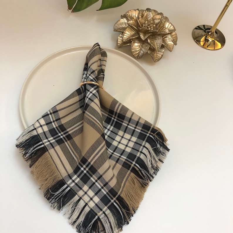 May include: A brown and black plaid napkin with fringed edges, folded and secured with a gold ring. The napkin is placed on a white plate.