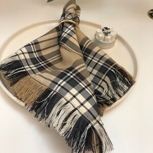 May include: A plaid fabric napkin with a fringed edge, folded and secured with a gold ring. The napkin is on a white plate with a small white candle holder.