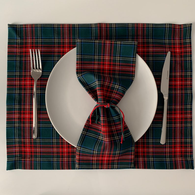 May include: A white dinner plate with a red and green plaid napkin folded into a bow on top. The plate is set on a red and green plaid placemat with a silver fork and knife on either side.