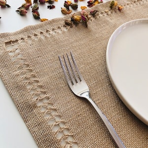 Puede incluir: Primer plano de un mantel individual de yute marrón con un borde de crochet blanco. Un tenedor de plata está sobre el mantel individual y un plato de cerámica blanco está en el lado derecho de la imagen.