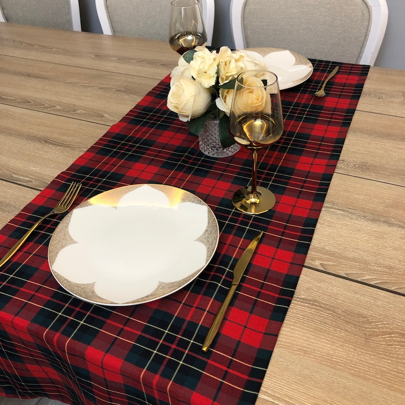 May include: Red and black tartan table runner with white flowers in a glass vase, two gold wine glasses, and a white plate with gold trim.