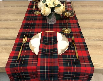 May include: Red and green plaid table runner with a white plate, gold silverware, and a gold wine glass. A bouquet of white flowers is in a clear glass vase on the table runner.