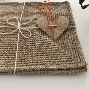 Puede incluir: Una estera cuadrada de crochet marrón con un corazón de arpillera y flores secas en la parte superior. La estera está atada con una cuerda blanca.