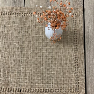 May include: A brown burlap table runner with a white lace trim. A small white vase with dried flowers sits on the runner.
