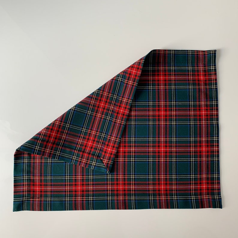 May include: A red and green plaid fabric placemat with a folded corner. The placemat is made of a woven fabric with a checkered pattern.