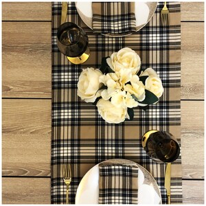 May include: A brown and black checked table runner with white accents. The runner is decorated with a bouquet of white flowers and two wine glasses. There are two gold forks and two white plates with matching checked napkins.