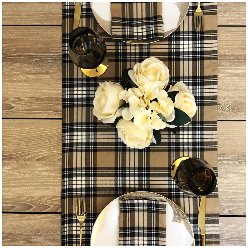 May include: A brown and black checked table runner with white accents. The runner is decorated with a bouquet of white flowers and two wine glasses. There are two gold forks and two white plates with matching checked napkins.