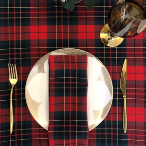 May include: A white dinner plate with a gold rim and a red and black tartan napkin on a red and black tartan tablecloth. A gold fork and knife are on the table.