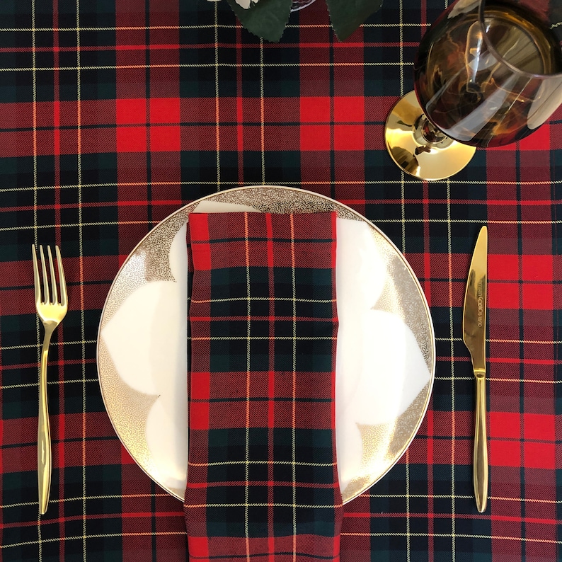 May include: A white dinner plate with a gold rim and a red and black tartan napkin on a red and black tartan tablecloth. A gold fork and knife are on the table.