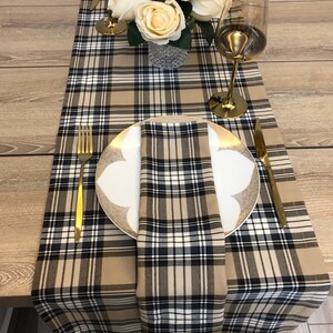 May include: A brown and black checked table runner with a white napkin folded on a white plate with a gold rim. A gold fork and knife are on the table.