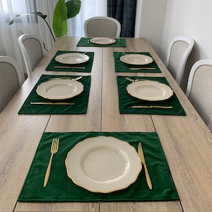 May include: A set of six green velvet placemats with gold trim, each with a white dinner plate and gold cutlery. The placemats are arranged on a light wood table.