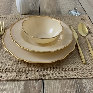 May include: A beige place setting with a gold rim on a brown hessian placemat. The place setting includes a bowl, a plate, a fork, a knife, and a spoon. All the utensils are gold.