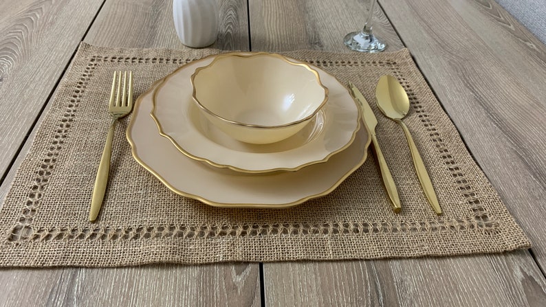 May include: A beige place setting with a gold rim on a brown hessian placemat. The place setting includes a bowl, a plate, a fork, a knife, and a spoon. All the utensils are gold.
