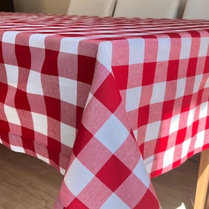 Puede incluir: Un mantel rectangular que presenta un patrón clásico de cuadros rojos y blancos. La tela muestra una cuadrícula consistente de cuadrados rojos y blancos alternos, drapeada sobre los bordes de una mesa para mostrar la textura y las esquinas.