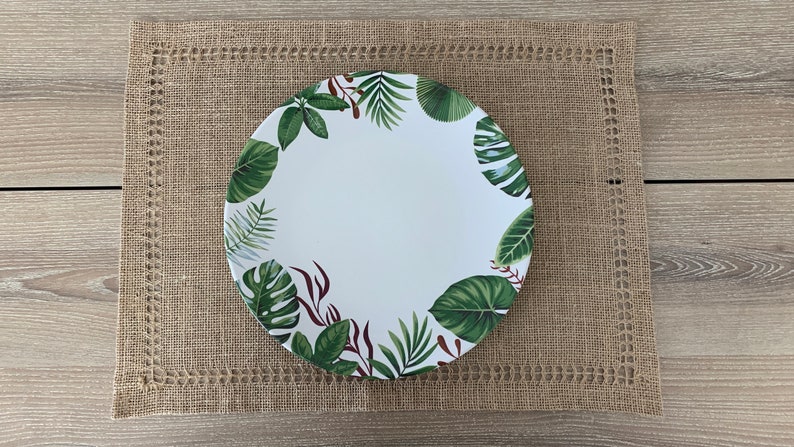 May include: A white ceramic dinner plate with a green and brown tropical leaf design. The plate is on a brown woven placemat.