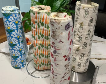 Op de afbeelding: Vier rollen papieren handdoeken met patronen. Ontwerpen omvatten witte madeliefjes op blauw, oranje wortelen, botanische illustraties en potten met bloemen. De rollen worden tentoongesteld op een wit aanrecht met een plant op de achtergrond.