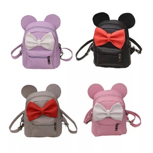 Puede incluir: Cuatro mini mochilas con detalles de lazo. Las mochilas son de color morado, negro, gris y rosa. Cada mochila tiene dos orejas pequeñas en la parte superior.