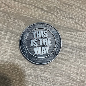 Op de afbeelding: Een zilverkleurige ronde munt met de tekst "THIS IS THE WAY" in hoofdletters. De munt heeft een gedetailleerd, gestructureerd ontwerp met een cirkelvormige rand en gestileerde vleugels.