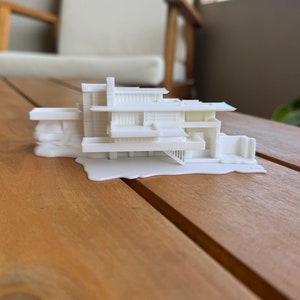 Może przedstawiać: Biały model domu w stylu nowoczesnym, wydrukowany w 3D, z płaskim dachem i wieloma poziomami. Model stoi na drewnianym stole.