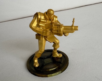 May include: A gold-painted figurine of a video game character holding a large gun. The character is standing on a round base with a stylised design.