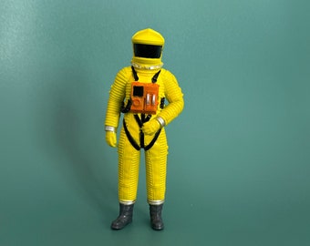 Könnte beinhalten: Eine gelbe Astronauten-Actionfigur mit schwarzem Visier und orangefarbenen Details. Die Figur trägt einen gelben Raumanzug mit schwarzen Trägern und einem schwarzen Gürtel.