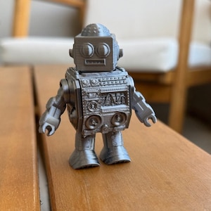 Könnte beinhalten: Ein silbernes Roboter-Spielzeug im Retro-Design. Der Roboter hat einen großen Kopf mit zwei runden Augen und einem kleinen Mund. Der Körper des Roboters ist aus Kunststoff und hat ein detailliertes Design. Der Roboter steht auf einem Holztisch.