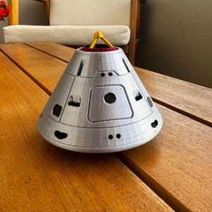 May include: A silver-colored, cone-shaped decorative object with black circular details and a yellow and red accent on top. The object is sitting on a wooden table. The design resembles a spacecraft or space capsule.