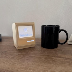 Puede incluir: Una unidad de pantalla en miniatura con forma de ordenador de color beige que muestra el logotipo de Apple y el texto "Think different." junto a una taza de café de cerámica negra. El dispositivo tiene una pequeña barra de luz horizontal.