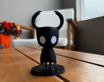 May include: A black figurine of a character with large curved horns and white eyes, standing on a circular base. The figurine is on a wooden table, with a blurred background of a white chair and plants.