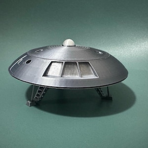 Puede incluir: Modelo plateado de platillo volante con parte superior abovedada y tres patas. El modelo tiene una pequeña cúpula redonda en la parte superior y ventanas rectangulares en la parte delantera. El platillo está sobre una superficie verde.