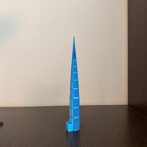 May include: A bright blue, three-dimensional model of a skyscraper featuring a tapered, pointed spire and a rectangular base. The structure displays a series of stacked, segmented levels that narrow towards the top, creating a sharp, vertical silhouette.