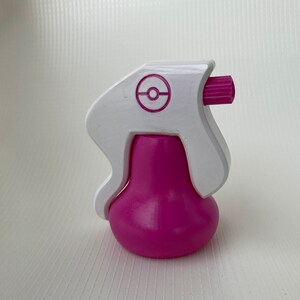 Puede incluir: Una botella de spray de pl&aacute;stico blanco con una base rosa y un logotipo de Pokeball en el lateral. La botella tiene una boquilla rosa.