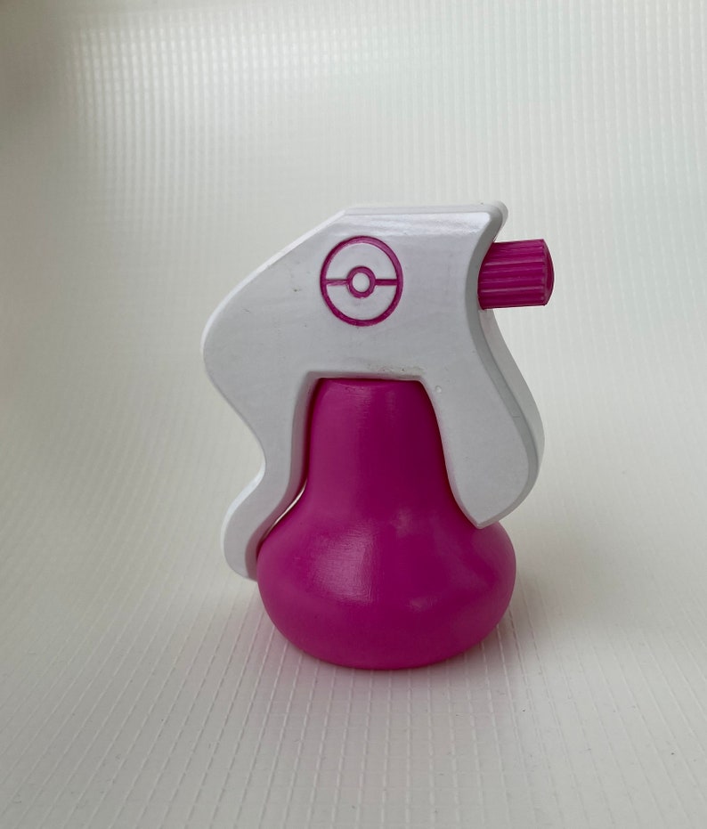 Puede incluir: Una botella de spray de pl&aacute;stico blanco con una base rosa y un logotipo de Pokeball en el lateral. La botella tiene una boquilla rosa.