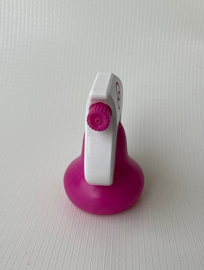Puede incluir: Una botella de spray de pl&aacute;stico rosa con un gatillo blanco y una perilla rosa. La botella tiene una base redonda y una etiqueta blanca con un c&iacute;rculo rosa.