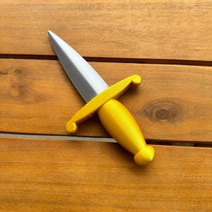 May include: A toy dagger featuring a metallic silver blade and a bright gold handle. The handle has a rounded grip and a crossguard with curved ends. The object is constructed with a smooth finish and a pointed tip, designed as a handheld prop.
