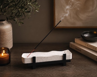 May include: A rectangular, off-white, textured stone incense burner tray with a wavy top edge, supported by a black metal frame. A single black incense stick with a red base is inserted into the tray, emitting a thin trail of white smoke.