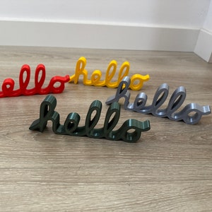 以下が含まれることがあります： 赤、黄、グレー、濃い緑の4つの3Dプリントされた「hello」のサイン。サインは木製の表面に一列に並んでいます。